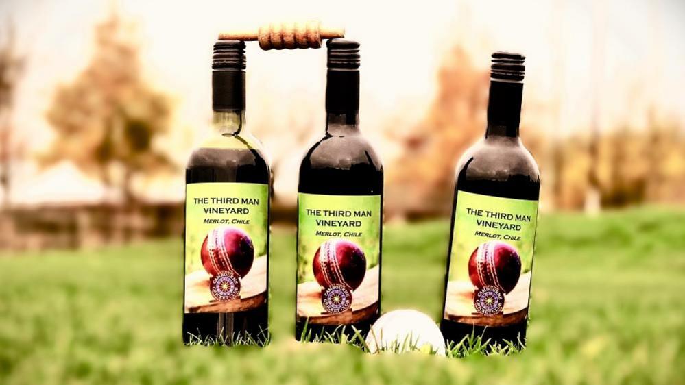 Three bottles of red wine set up like cricket stumps on grass. A bail sits between the top of left hand had middle bottles. The right hand bottle is leaning to the right, A white cricket ball sits on the grass between the middle and right-hand bottles.