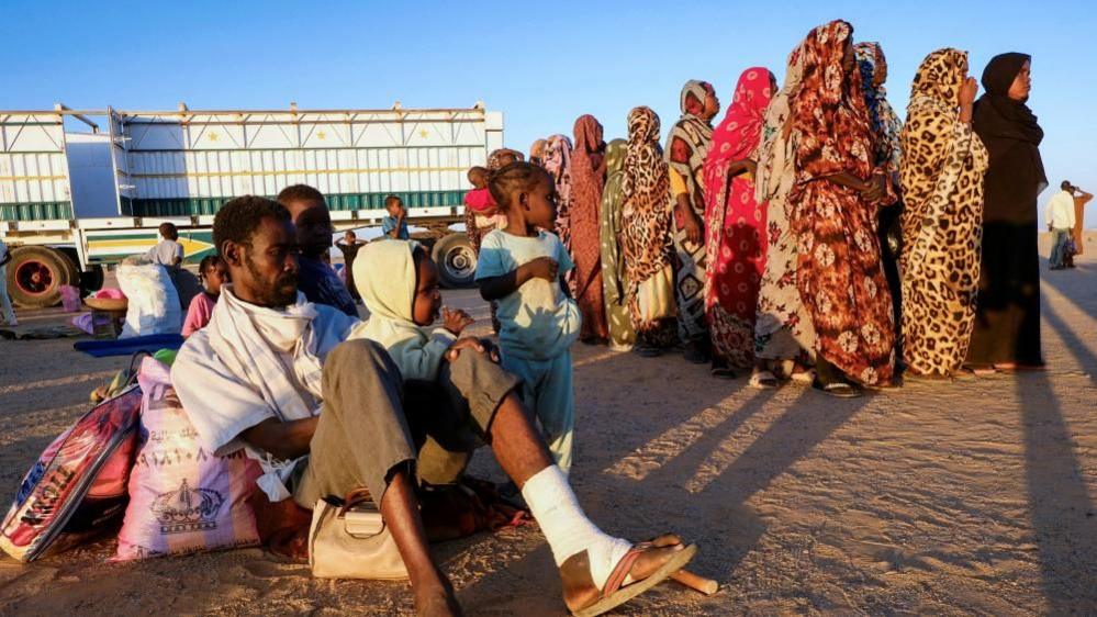 An injured man from el-Fasher sits on the ground surrounded by his children. A queue of women can be seen on the right of the picture and and empty open-bed lorry is in the background.