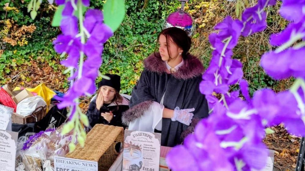 A young boy in a black hat sits on a chair and a girl in clock with a fir collar stands at a market stall selling sweets. in the foreground are purple flowers hanging from the stall.