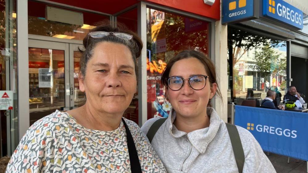 Paula Patterson is wearing a floral patterned top, and she has her glasses up on her hair, which is dark coloured and tied back. She has a bag strap over her left shoulder. Her daughter Demi Stroud is wearing a grey top with a zipped front, and has rucksack straps over her shoulders. She has long brown hair, tied back, and is wearing glasses.