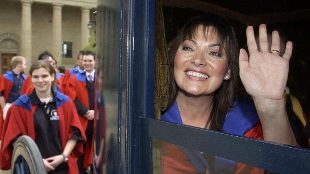 Lorraine Kelly looks out of a carriage window and waves. She is wearing red and blue robes. People stand behind the carriage also wearing red and blue robes.