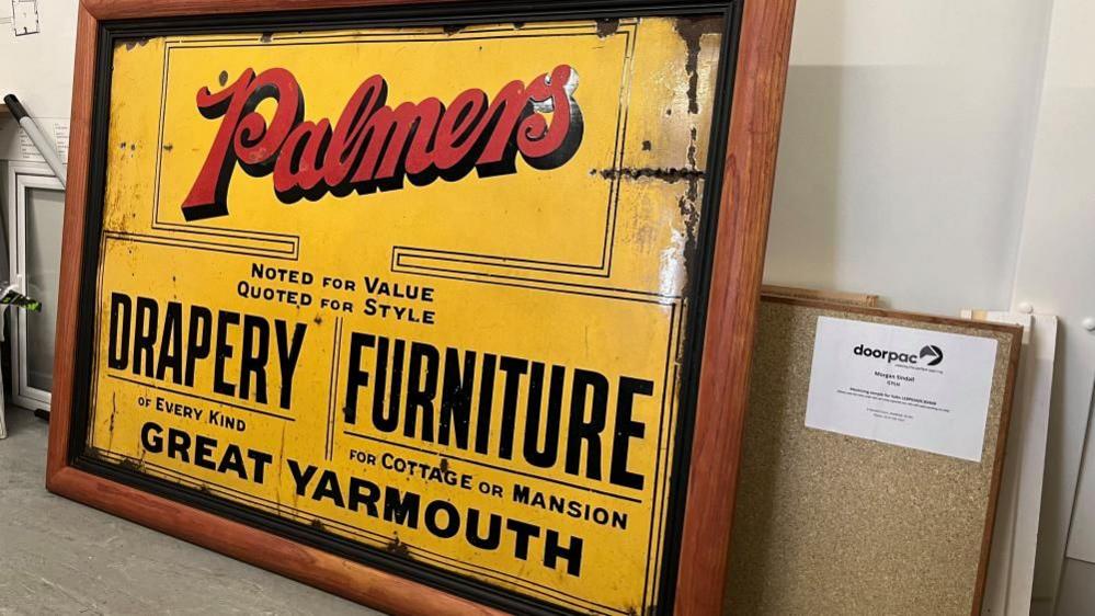 Yellow enamel sign, in a wooden frame, with black text, stating:
PALMERS
NOTED FOR VALUE; QUOTED FOR STYLE
DRAPERY OF EVERY KIND
FURNITURE FOR COTTAGE OR MANSION
GREAT YARMOUTH
