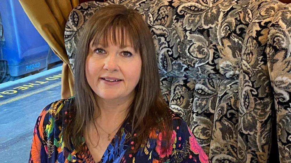 Councillor Kerry Robinson Payne, wearing a multicoloured blouse, sitting on a luxurious train. She is smiling, and has long brunette hair. The seat fabric is highly patterned.