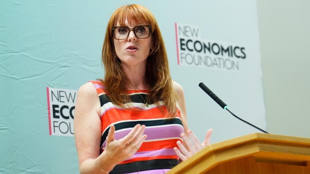 Angela Rayner is stood up and speaking at a wooden lectern. She is wearing a dress with multi-coloured horizontal stripes. She is wearing glasses and has long, ginger-coloured hair. Rayner is stood in front of backdrop which has "New Economics Foundation" written on it.