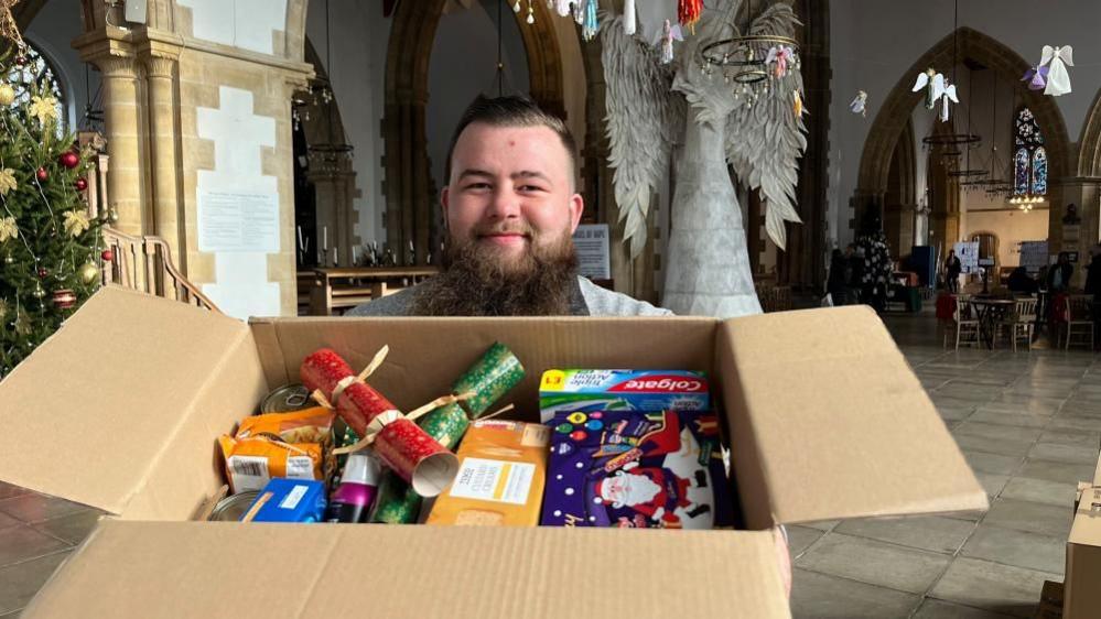 John McBride, holding a Christmas hamper box, filled with food and Christmas crackers. He is standing in Great Yarmouth Minster, where a giant model of an angel stands in the background.