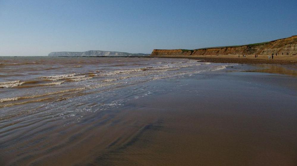 Brook Bay on the Isle of Wight. Calm waves lap onto the sandy beach, cliffs are in the distance.