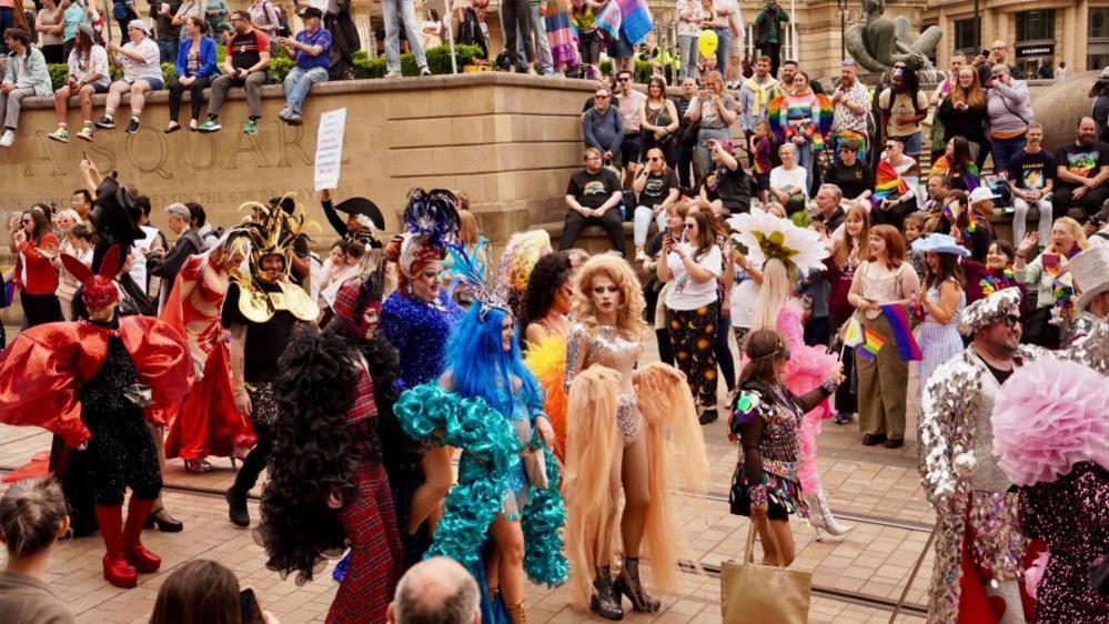 A Pride parade makes it way through a city centre. Drag queens are walking through the middle of the street which is lined with crowds. Lots of people are waving Pride flags. 