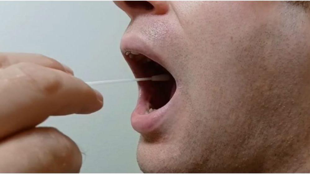 The side profile of a man's face with his mouth open as he does a lateral flow test