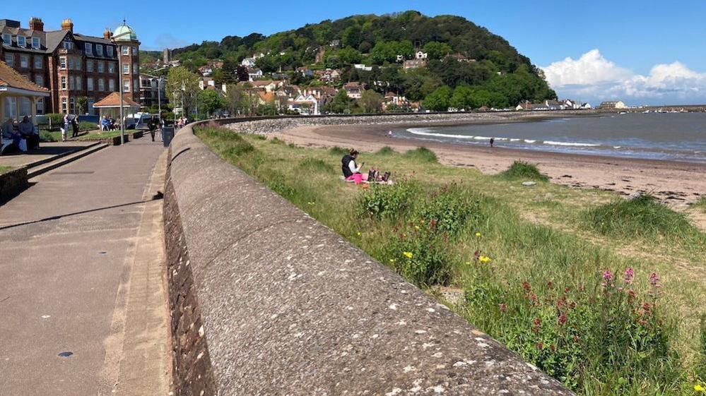 A promenade in Minehead, with the sea at high tide on the right, while North Hill sits in the background. 