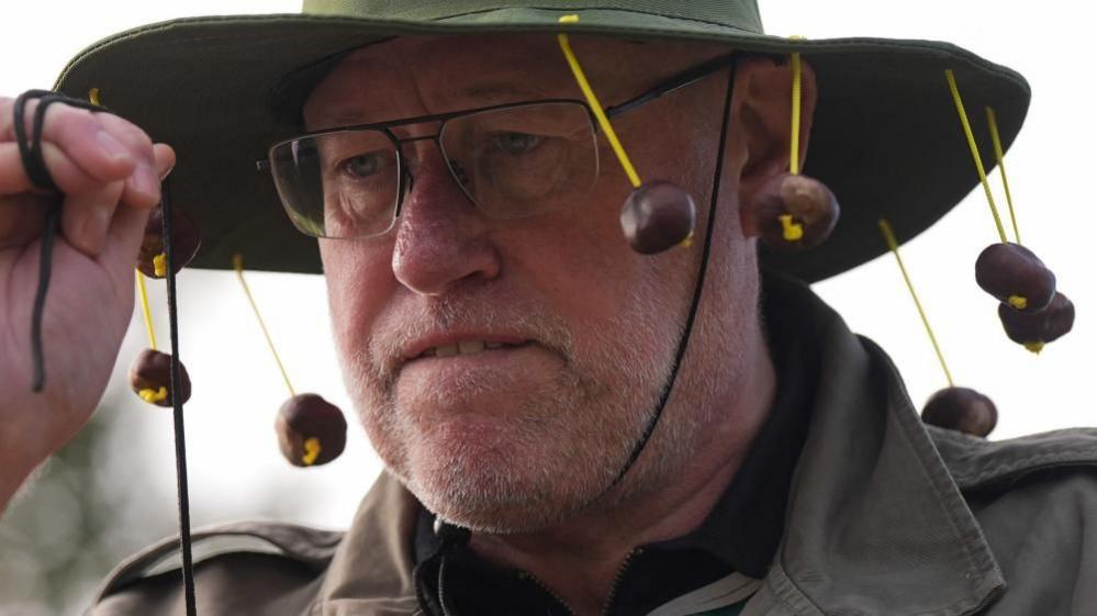 A man with glasses wearing a wide brimmed hat with conkers dangling from it