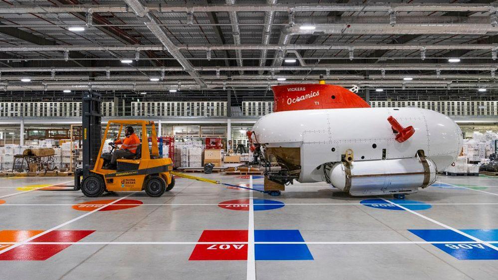 A forklift truck pulling a submarine across a grey floor with painted grid. The sub is rounded and white with a red fin on top.