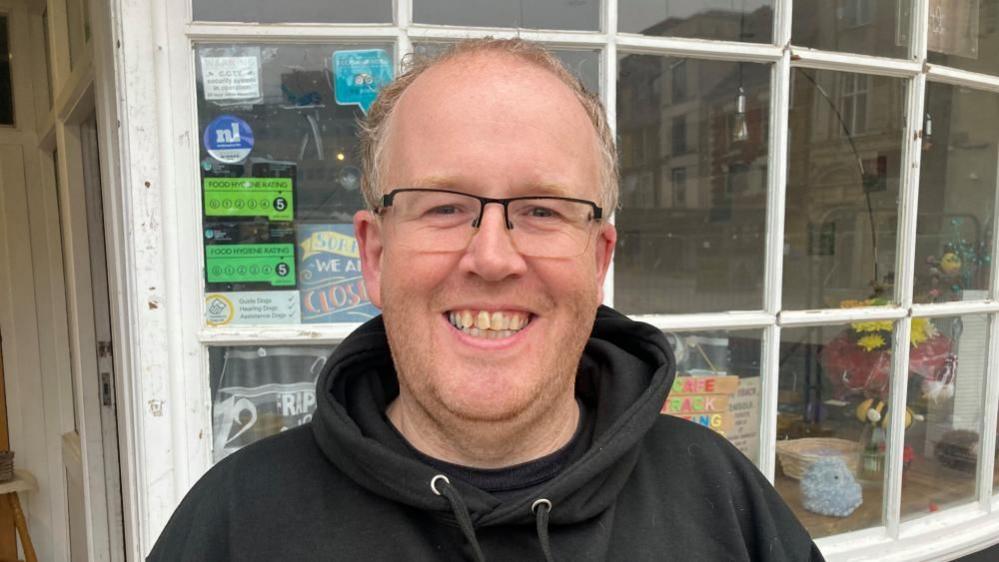 Tom Cliffe with short light-coloured hair wearing glasses and a black fleece, standing outside Cafe Track where flowers and hygiene ratings are visible in small windows.