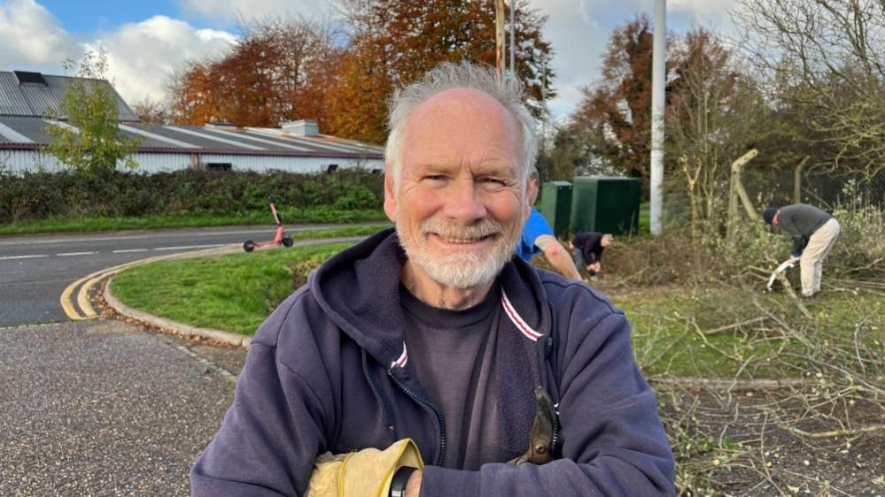 Tony Heath with a navy blue top, grey hair and a beard smiling. Behind him are three men clearing some trees.