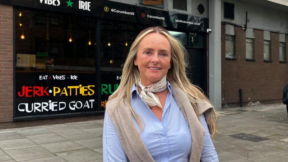 Amanda Massey with long blonde hair, wearing a blue shirt and white scarf while smiling at the camera in front of a restaurant with words including JERK, PATTIES and CURRIED GOAT painted on the outside.