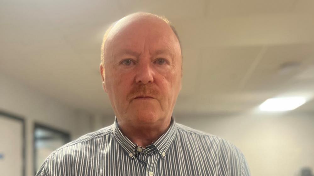 A bald man looking at the camera. He has a moustache and is wearing a black and white striped shirt. 
