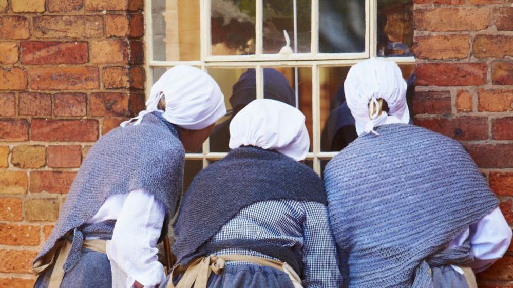 Three women in Victorian poorhouse costume, looking through a large workhouse window