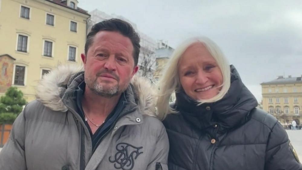 Mr Fenton with short dark hair wearing a grey anorak with white hood standing next to Mrs Fenton, who has long blond hair, wearing a dark-coloured anorak. She is smiling. They are in a square in Poland - a historic building is visible in the background.