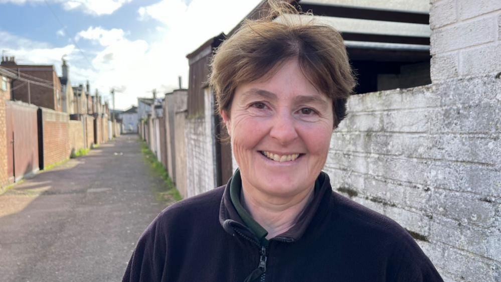 Kerry Hemsworth, standing in an alleyway cleared of rubbish and bins. She has auburn hair and is wearing a black fleece jacket.