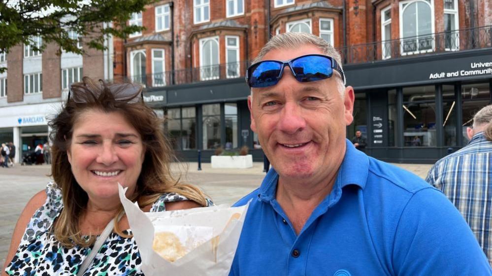 Susie and Kenny Hearn, sitting in Great Yarmouth Market Place. She is wearing a peacock-tail patterned top, and has long brunette hair with her sunglasses on her head. Kenny is wearing a blue polo shirt, and has tinted sunglasses on his head. He is holding a cone of chips and gravy. Behind them is The Place, the new library and learning centre in Great Yarmouth Market Place.