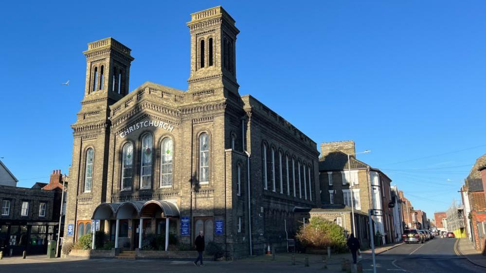 Christchurch church in Great Yarmouth, a large brick building with a gabled end and large windows. Two brick towers adorn the building. There is a blue sky behind it.