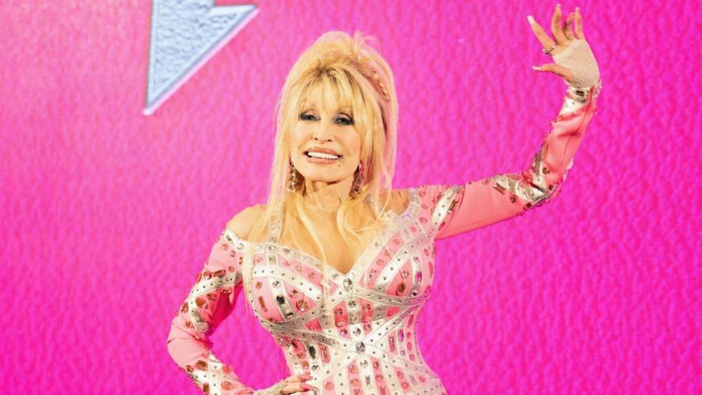 Dolly Parton, standing in front of a pink background, raises her left arm as she waves, while her other arm is on her hip.