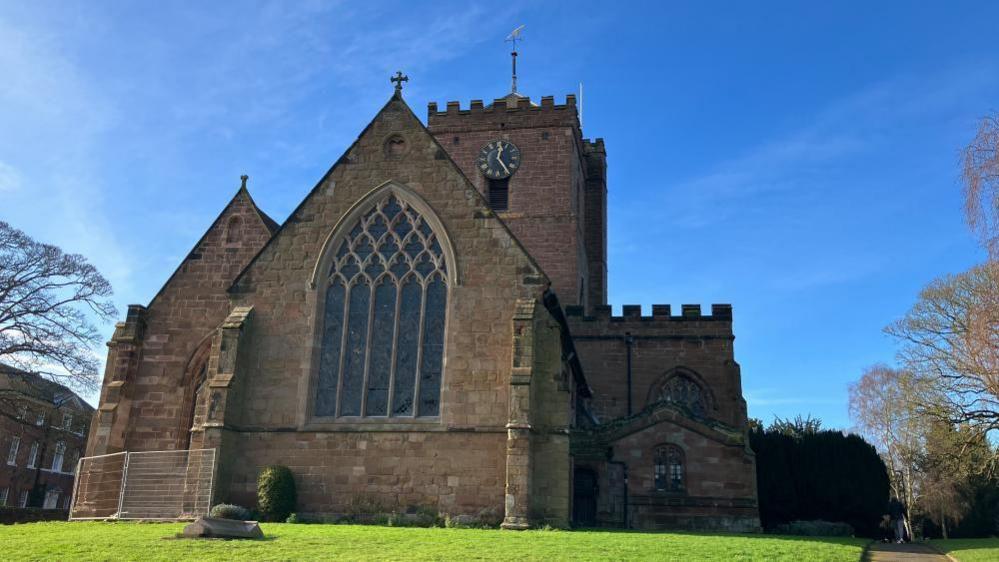 Bright blue sky frames the background, with a stone church at the front of the picture and grass along the bottom. A few trees to either side of the church. 