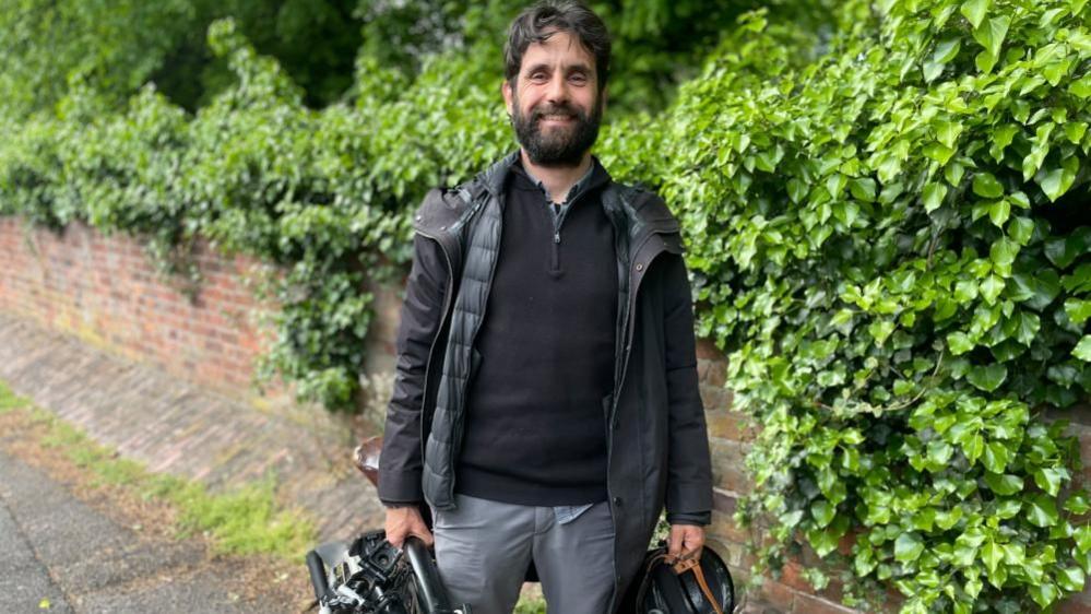 A portrait image of Miguel stood on a pavement by the QMC roundabout. He is holding a folded bicycle in one hand and a helmet in the other.