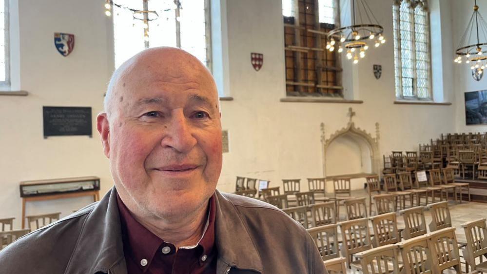 A man standing inside a church, where the window to the right of him is boarded up. He is clean shaven, bald and wearing a burgundy shirt, with a grey/brown jacket over it. There are chairs placed and stacked behind him, and hanging chandeliers, which are lit.
