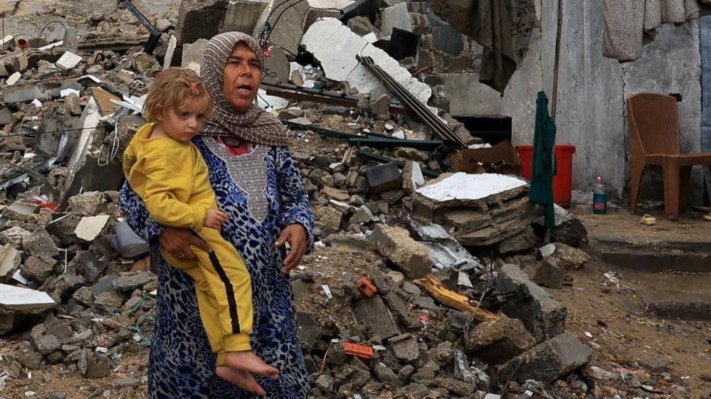 A Palestinian woman holds a child dressed in a yellow track suit, with a pile of rubble behind them.