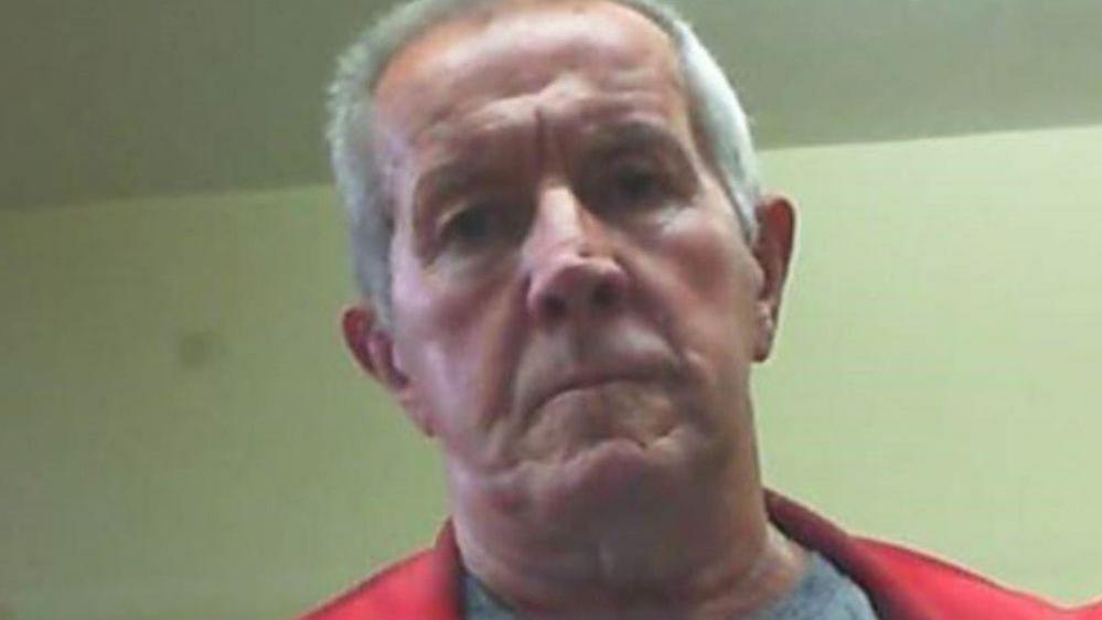 Richard Scatchard is wearing a red jacket and is looking down at the camera