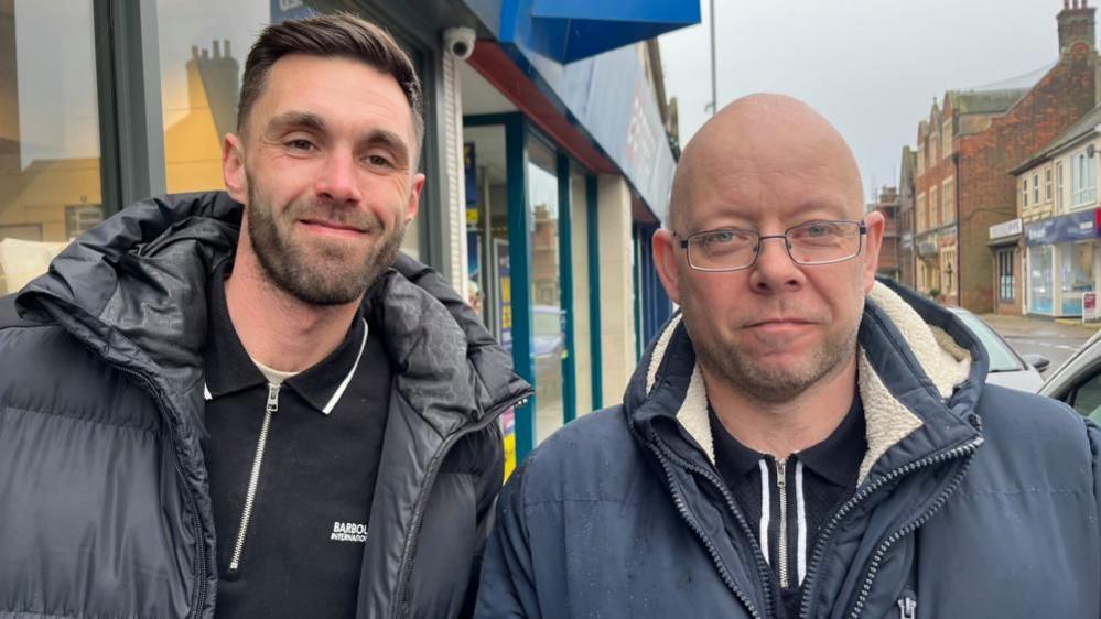 Dale Nichols is on the left, with brown hair swept to his right side. He has a beard and a moustache. He is wearing a black padded jacket and a black zip-fronted polo shirt and he is smiling. Mark Williams, on the right, is bald and wears glasses. He is wearing a blue jacket with faux fur white lining and a zipped-up polo shirt.