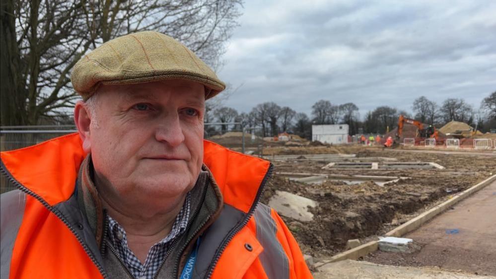 Alistair Beales, wearing a tweed flat cap, orange high-visibility jacket and checked shirt. He is standing next to a construction site.