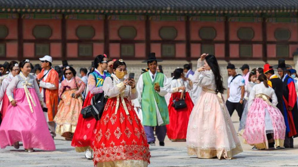 Dozens of tourists wearing traditional Korean clothes - called hanbok - are visiting Gyeongbokgung Palace in Seoul. A section of the Gyeongbokgung Palace - a row of doors - can be seen behind the groups of people in colourful clothing.