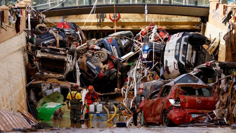 At the entrance to a road tunnel, two firefighters operate a pump. Behind them, the entrance is entirely blocked by dirty, damaged cars haphazardly piled up.