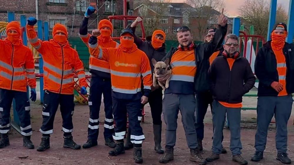 A group of men wearing high-vis orange workwear and orange balaclavas stand with their fists raised in the air on a playground.