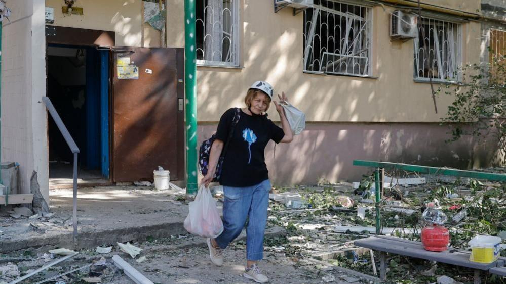 A woman wearing a black top, jeans, trainers and a hard hat, and holding two plastic bags in both hands emerges out of a house after a Russian attack. Debris can be seen all around.