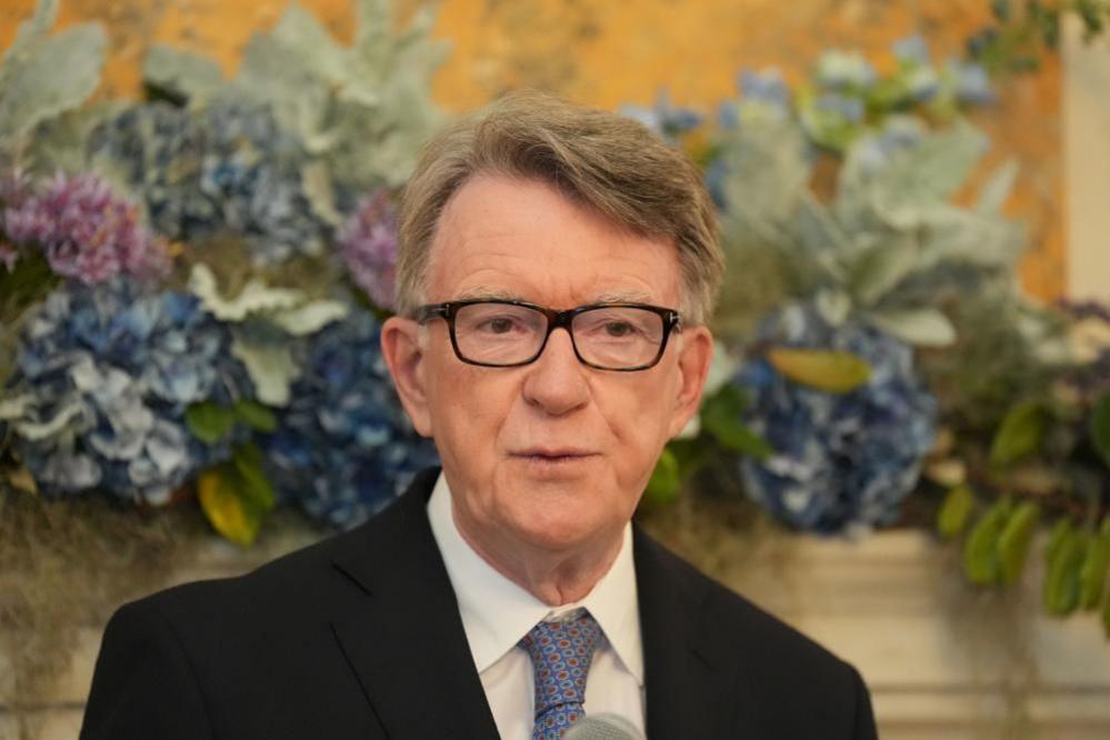 Peter Mandelson, wearing black framed glasses, a black suit, shite shirt, and blue tie