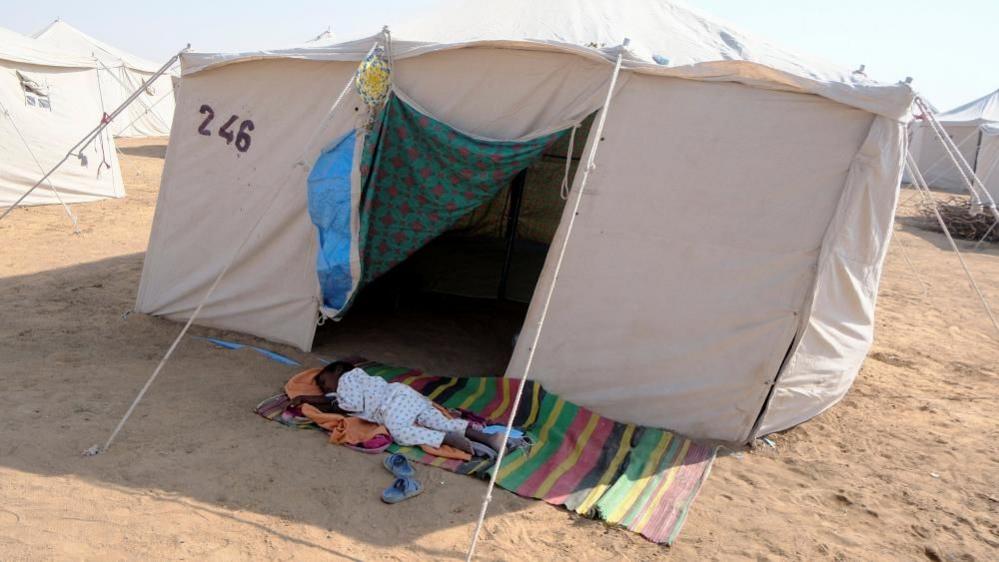 A child from el-Fasher sleeps on the ground on a rug outside a white tent near the displacement camp in al-Dabbah, Sudan, 12 November 2025.