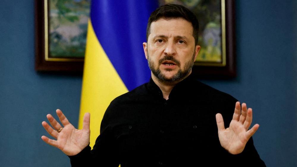 President Zelensky in his trademark black top holding his hands up in front of a Ukrainian glad and two paintings on a dark blue wall