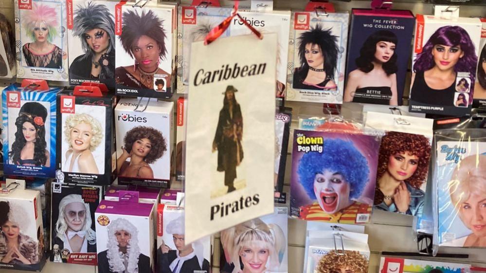 A sign saying "Caribbean Pirates" hanging in front of boxes of wigs. The boxes show images of men and women with various hair styles. One box advertises a Marilyn Monroe wig.