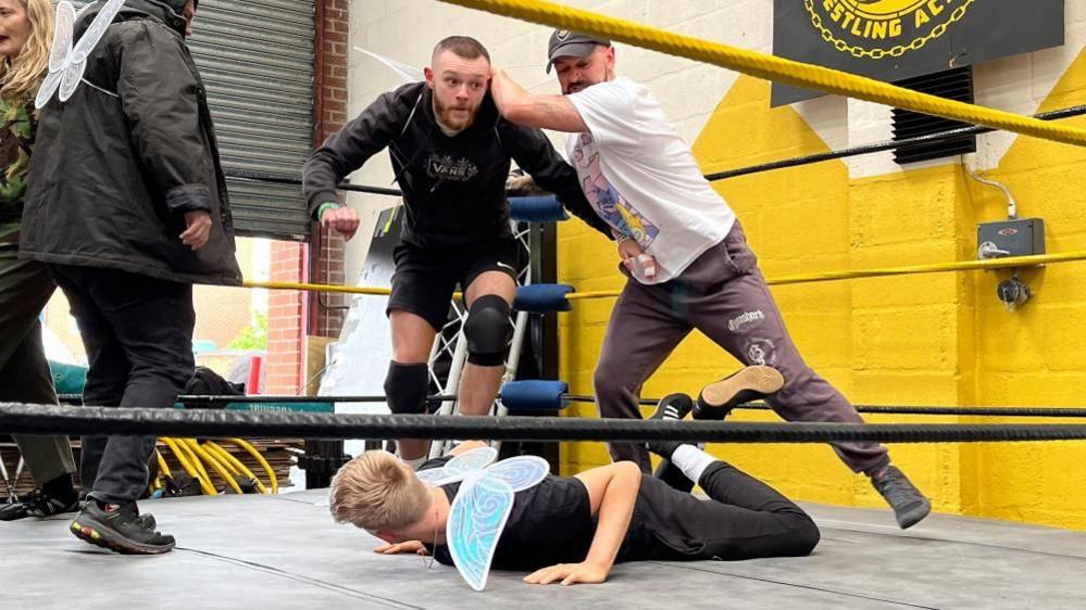 The character Nick Bottom preparing to throw an unnamed fairy actor to the ground, in a two on one wrestling battle.