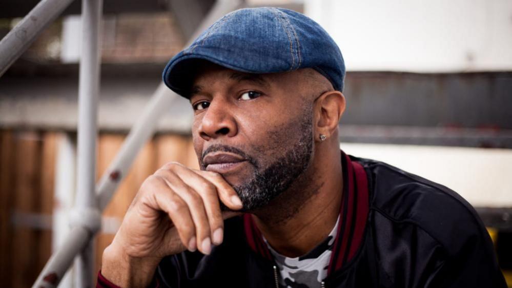 Raymond Stevenson, with a shaped beard and moustache, sits with his right hand against his chin. He is looking at the camera, wearing a blue denim flat cap, and a black, white and grey camouflage tee-shirt with a black jacket, featuring a red and black knitted collar, over.
There is a staircase bannister visible to the left of the image, and a building out of focus, to the backdrop.