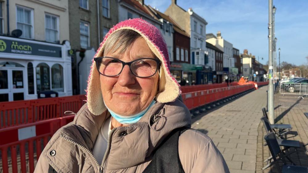 Susan Bayfield is wearing black rimmed glasses, and has a blue face mask around her neck. She is wearing a pink felted hat and has a beige coat over a cream-coloured top. She is standing in the market place in Great Yarmouth, where there is orange-coloured barriers running through during paving works.