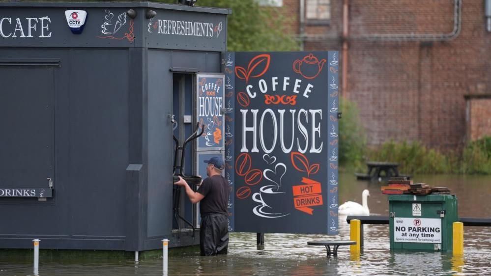 A grey wooden building with "Coffee House" signs has floodwater around the base. A man is removing a wooden table from the building while standing in the water. A swan swims by in the background.