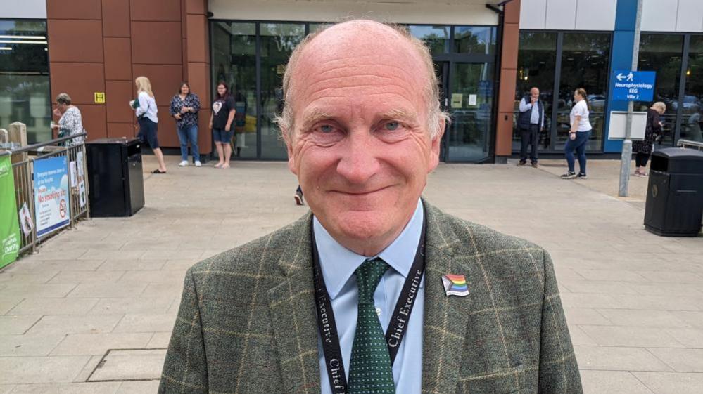 Nick Hulme is standing in front of Colchester hospital  in green checked jacket with rainbow pin and “Chief Executive” lanyard. He is smiling at the camera. 