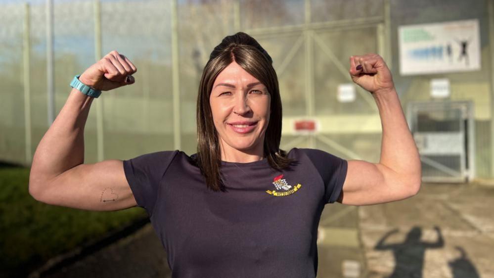 A woman with brown hair flexes her biceps to display her muscular arms. She wears a blue t-shirt with a 'Fit to Serve' badge and stands in a prison yard.