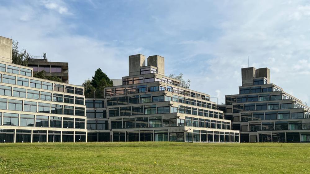 Ziggurat buildings at the University of East Anglia. There are six stories of windows, with three buildings shown. Green grass can be seen at the bottom of the image, and above the buildings the sky is blue with a few wispy clouds. 