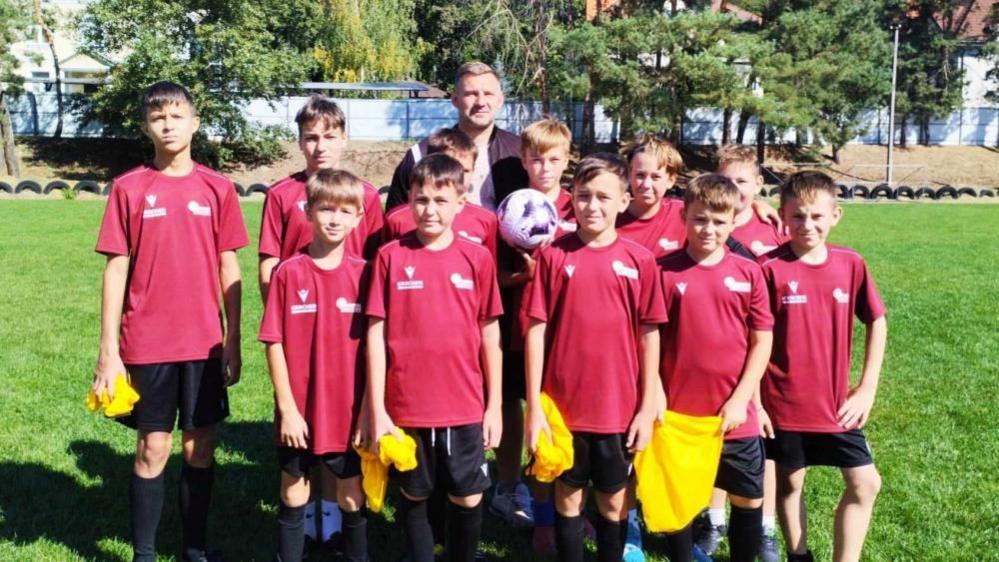 Boys wearing red football shirts and a coach line up around a white and purple football on a grass pitch