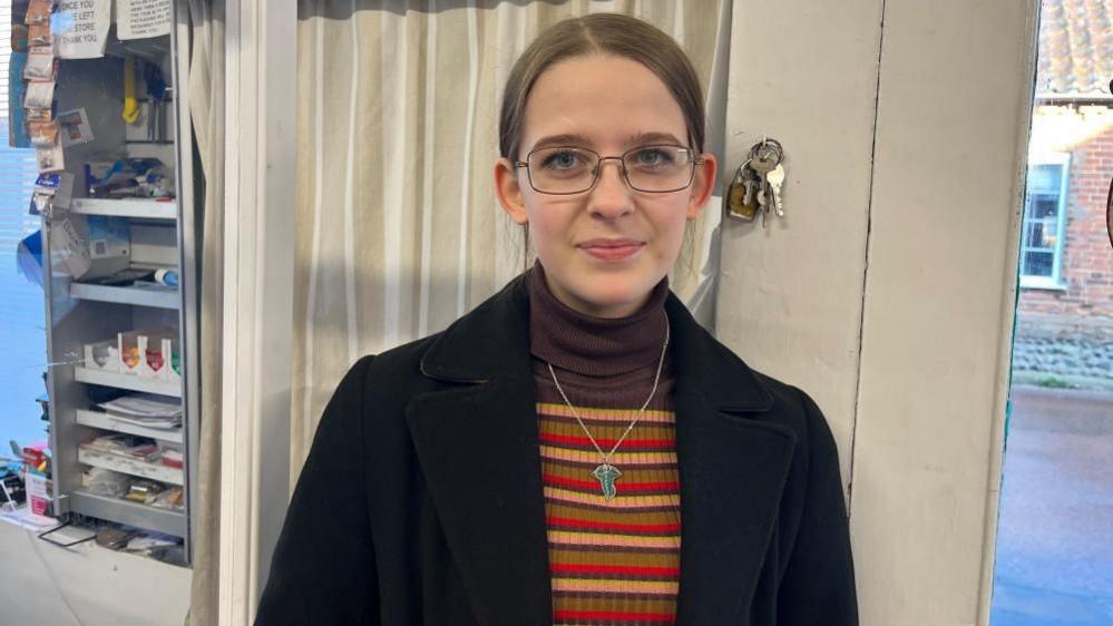 Kami Matthew, standing in a shop, by a door, with a set of keys behind her head. She is wearing a necklace, a stripped top and black coat. She is looking straight at the camera. 