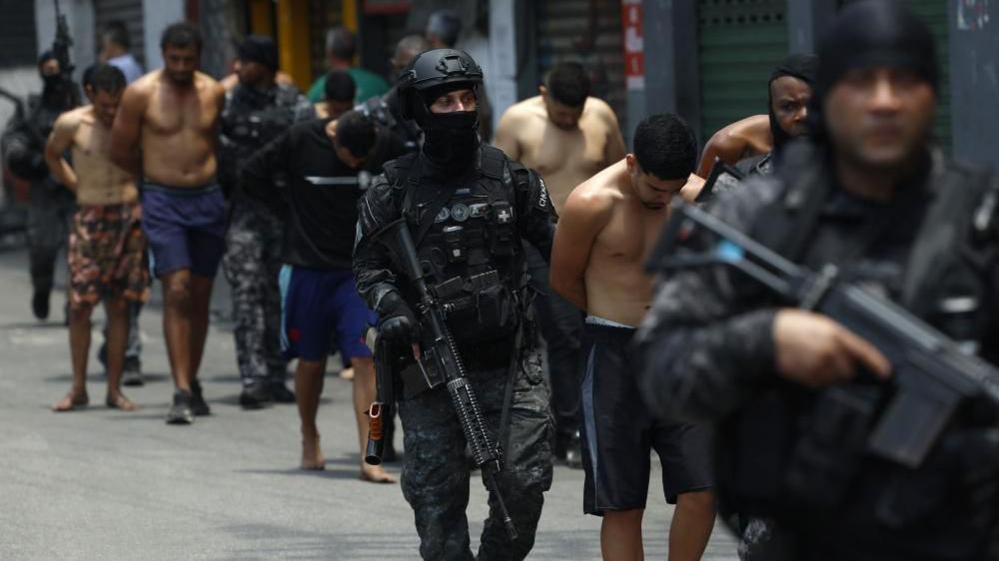 Rio de Janeiro Police officers guard a group of people during an operation in Rio de Janeiro. The officers are carrying weapons and one of them is wearing a balaclava. The men they are escorting have been handcuffed.